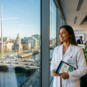Uma farmacêutica brasileira profissional em um jaleco branco olha com confiança por uma grande janela de escritório com vista para o centro de Dublin, na Irlanda, incluindo o rio Liffey e Trinity College. Ela segura um tablet com dados moleculares. O escritório tem a marca da Global Agency.